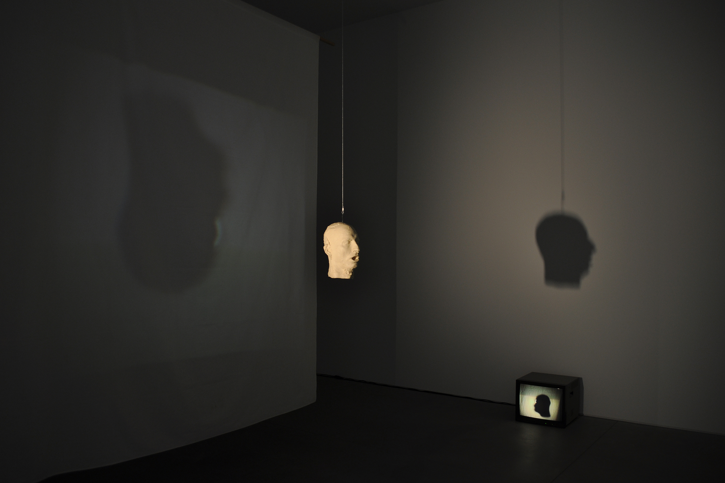 A white figure head is suspended from the ceiling in a black room. The shadow of the head plays on the wall.
